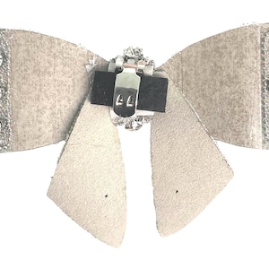 May include: A white faux leather bow with a silver rhinestone embellishment and a silver metal clasp. The bow is designed to be attached to clothing or hair.