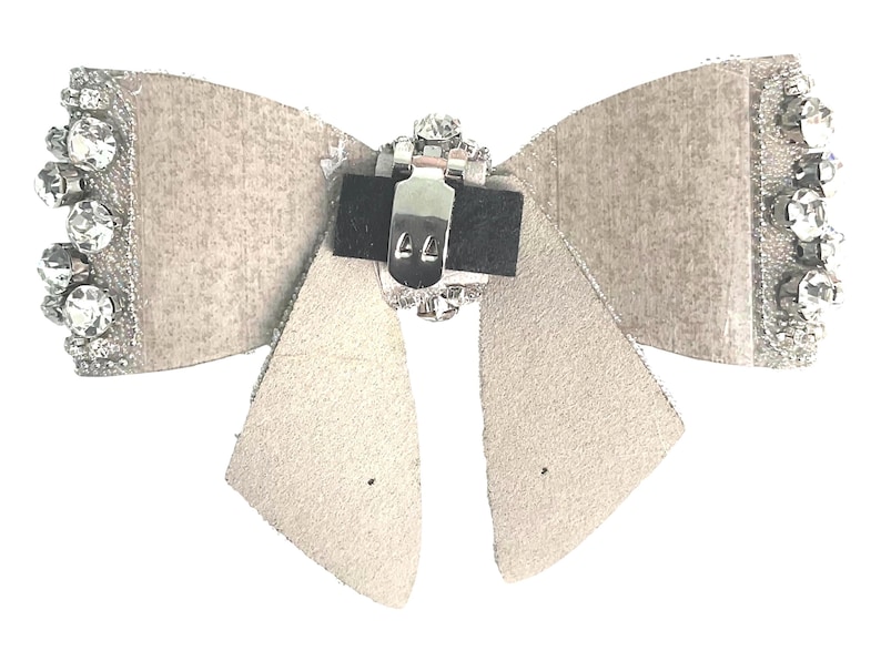 May include: A white faux leather bow with a silver rhinestone embellishment and a silver metal clasp. The bow is designed to be attached to clothing or hair.