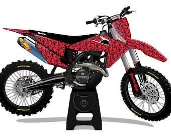 May include: A red and black motocross bike with a patterned design. The bike has black tires, a silver exhaust, and is supported by a black stand. The bike has the text "Graphics Guys" on the front suspension.