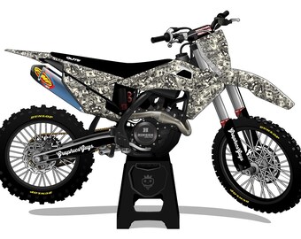 May include: A motocross bike with a money-themed graphic design. The bike features black tires, a black seat, and a blue exhaust. The text "Graphics Guys" is visible on the frame. The bike is on a black stand.
