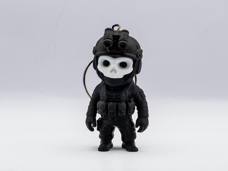 May include: A black figurine of a soldier wearing a helmet with night vision goggles and a skull-shaped mask. The soldier is dressed in tactical gear and has a keychain attachment. The figurine is set against a white background.