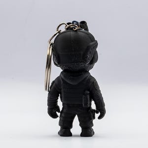 May include: A black keychain featuring a figurine of a soldier in tactical gear. The figurine has a helmet, vest, and utility pouches. The keychain has a silver ring attached to the top of the figurine.