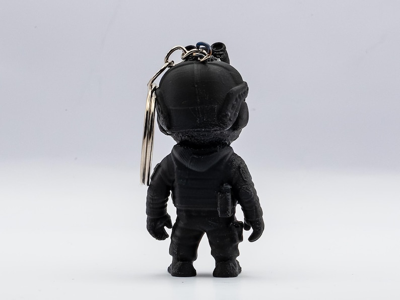 May include: A black keychain featuring a figurine of a soldier in tactical gear. The figurine has a helmet, vest, and utility pouches. The keychain has a silver ring attached to the top of the figurine.