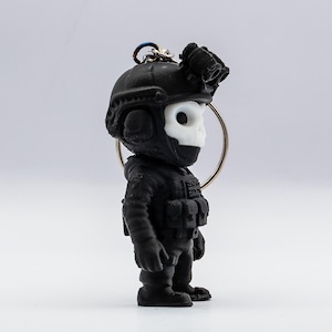 May include: Black keychain featuring a stylized soldier figure with a skull-shaped face. The figure wears a tactical helmet with night vision goggles and a tactical vest. The keychain is attached to a silver ring.