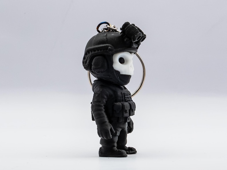 May include: Black keychain featuring a stylized soldier figure with a skull-shaped face. The figure wears a tactical helmet with night vision goggles and a tactical vest. The keychain is attached to a silver ring.