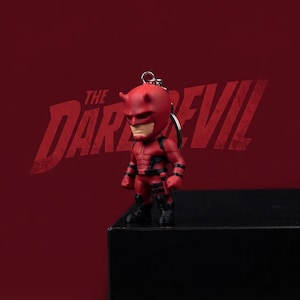 May include: A red and black Daredevil character keychain featuring a stylized figure with a mask, horns, and a muscular suit. The figure is attached to a silver metal keyring. The background displays the text THE DAREDEVIL in large red letters.