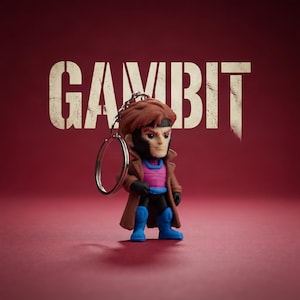 May include: A Gambit keyring featuring a stylised figurine with brown hair, a brown trench coat, a magenta chest piece, black gloves, and blue boots. The word GAMBIT is printed in large, distressed white capital letters in the background.