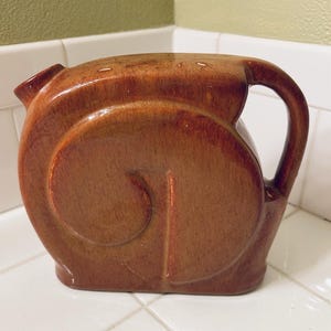 May include: A brown ceramic pitcher with a rounded body and a handle. The pitcher has a spout and a decorative swirl design on the front. The pitcher is sitting on a white tiled surface.
