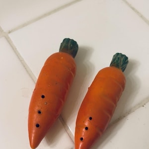 May include: Two orange carrot-shaped ceramic whistles with green tops and three holes each. The whistles are on a white tiled surface. The carrots are a bright orange color.