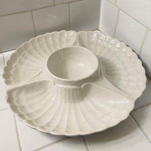 May include: Off-white ceramic serving dish with a central dip bowl and three surrounding compartments. The dish has a scalloped, fan-like design. Ideal for serving appetizers or snacks.