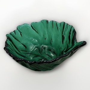 Op de afbeelding: Een groene glazen kom in de vorm van een blad. De kom heeft een getextureerd oppervlak met donkergroene accenten en een zwarte rand. De bladvormige kom is een decoratief item.