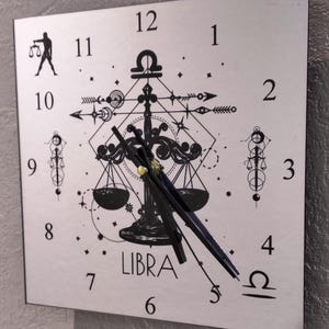 Könnte beinhalten: Eine weiße Uhr mit schwarzen Zeigern und einem schwarzen Sternzeichen Waage in der Mitte. Das Zifferblatt hat schwarze römische Ziffern und ein schwarzes Waage-Symbol unten.