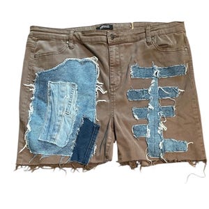 May include: Brown denim shorts with distressed patches of light and dark blue denim. The shorts have a frayed hem and a button closure. The patches are sewn on with white thread.