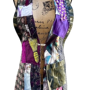 May include: A sleeveless vest constructed from a patchwork of various fabrics in shades of purple, green, black, and white. The garment features a mix of geometric, floral, and camouflage patterns, secured at the center with a single silver safety pin.