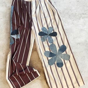 May include: Wide-leg pants with a unique design. One leg features vertical burgundy stripes with white lines, while the other has white stripes with burgundy lines. Denim flower appliqués add a decorative touch. The pants are made of a lightweight fabric.