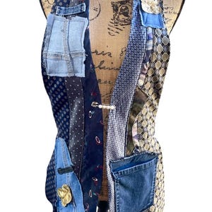 May include: A sleeveless vest constructed from various repurposed neckties and denim fabric patches. The garment features a mix of blue, grey, and gold patterns, including polka dots and geometric shapes, with a pearl-accented safety pin closure.