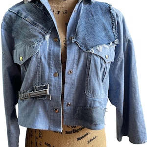 May include: A light blue denim jacket with patchwork detailing. The jacket features a collared neckline, button-down closure, and multiple pockets. Patches of darker denim are sewn onto the shoulders, chest, and lower front. The jacket has long sleeves.