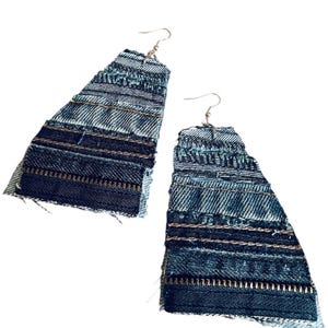 May include: A pair of handmade denim earrings. Each earring is a layered, triangular shape with varying shades of blue denim. The design incorporates visible stitching and zipper details, creating a unique, textured accessory.