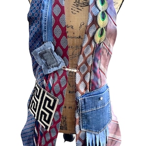 May include: A sleeveless vest constructed from various neckties and denim fabric patches. The garment features a mix of blue, burgundy, and brown patterned silk ties, a denim pocket, a Greek key motif patch, and a decorative pearl safety pin.