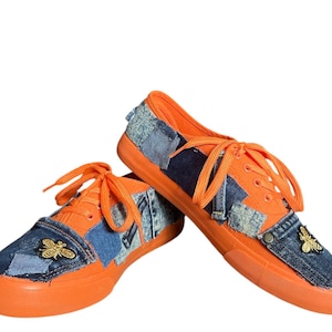 May include: A pair of orange sneakers with denim patchwork and orange laces. Each shoe features a gold bee embellishment on the denim. The soles and the tongue of the shoes are orange.