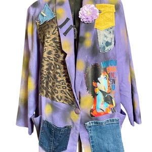 May include: A purple blazer featuring yellow spray-painted spots, a lavender fabric flower, and various patches including leopard print, denim, and a graphic print of a person with the text "HEY YOU GINA N, N". It has a single button closure.
