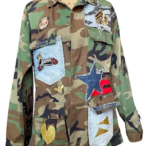 May include: Camouflage print jacket with denim patches and decorative embellishments. Features a large blue star, gold glitter heart, and patches with text and symbols. The jacket has a button-up front and multiple pockets.