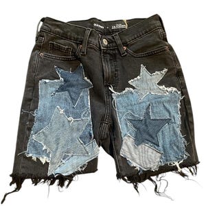 May include: Black denim shorts with frayed edges and patches of light and dark blue denim stars. The shorts have a classic five-pocket design and the brand name "OLD NAVY" is visible on the waistband.