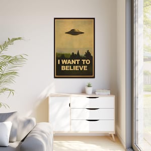 May include: A framed poster with a vintage aesthetic, featuring a flying saucer above a landscape of trees. The text "I WANT TO BELIEVE" is displayed in bold, gold lettering. The poster is displayed on a white cabinet.