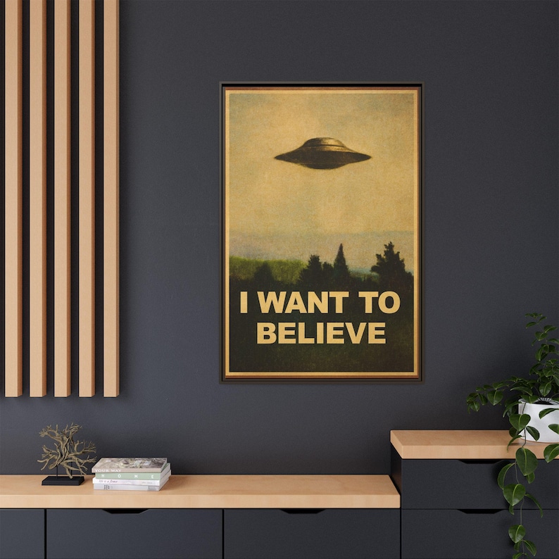 May include: Framed poster featuring a vintage-style image of a UFO hovering above a forest. The text "I WANT TO BELIEVE" is printed in bold, gold letters below the image. The poster is displayed on a dark blue wall.