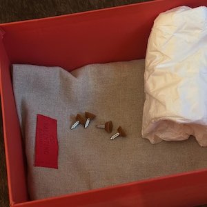 Puede incluir: Una caja roja abierta que contiene una tela gris, peque&ntilde;os tachuelas marrones y plateadas y un fajo de papel de seda blanco. Una etiqueta roja con la marca "Valentino Garavani" est&aacute; adherida a la tela.