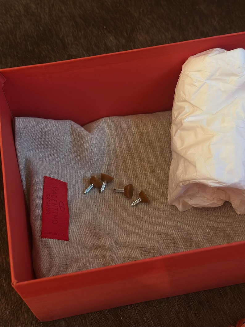 Puede incluir: Una caja roja abierta que contiene una tela gris, peque&ntilde;os tachuelas marrones y plateadas y un fajo de papel de seda blanco. Una etiqueta roja con la marca "Valentino Garavani" est&aacute; adherida a la tela.