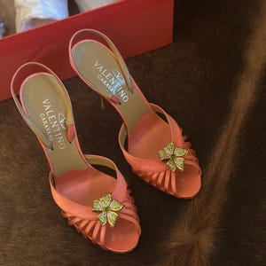 Puede incluir: Un par de zapatos de tac&oacute;n con correa trasera Valentino Caravan de color coral. Los zapatos presentan un adorno floral con detalles verdes y dorados. Los tacones est&aacute;n sobre una superficie marr&oacute;n, y la caja es visible en el fondo.