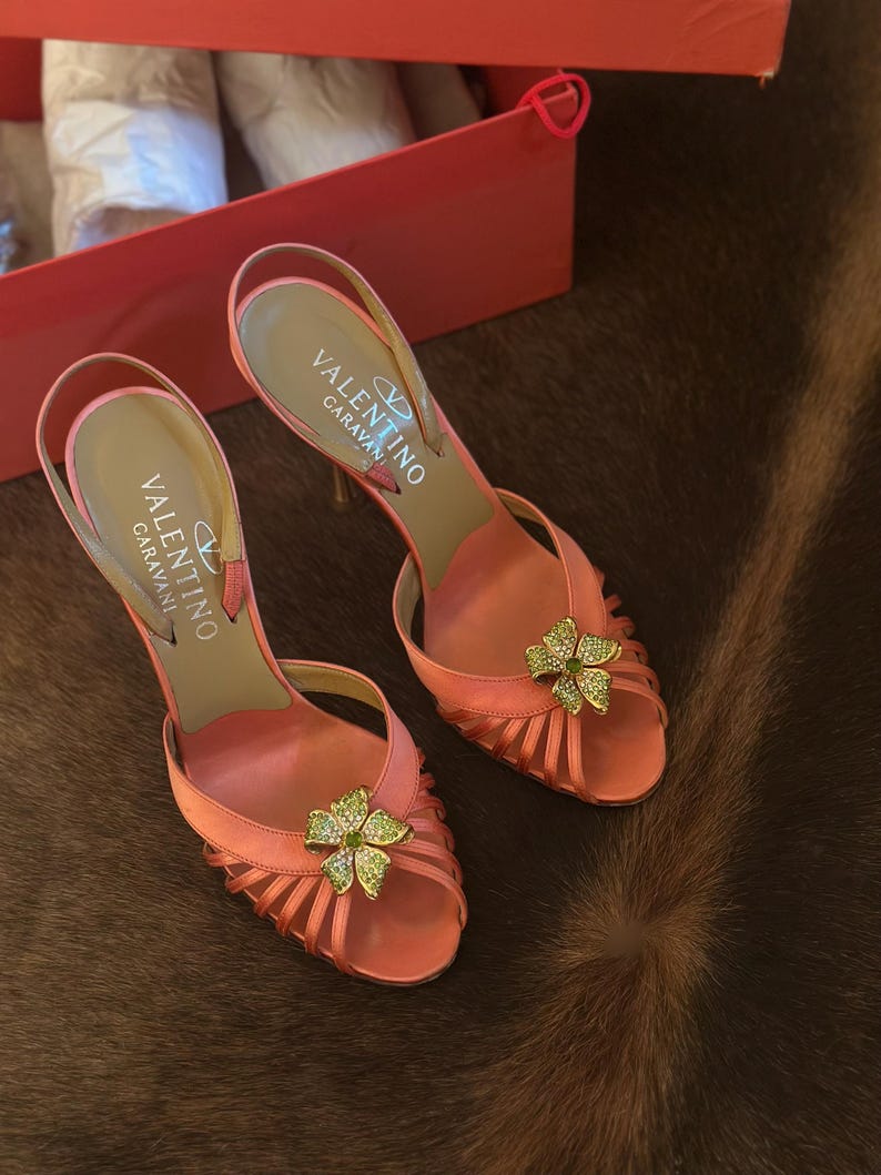 Puede incluir: Un par de zapatos de tac&oacute;n con correa trasera Valentino Caravan de color coral. Los zapatos presentan un adorno floral con detalles verdes y dorados. Los tacones est&aacute;n sobre una superficie marr&oacute;n, y la caja es visible en el fondo.