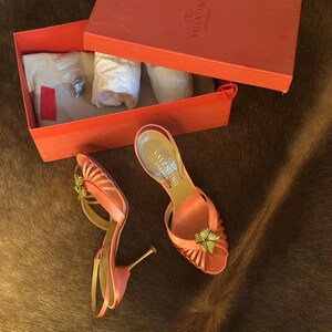 Puede incluir: Un par de sandalias de tac&oacute;n alto Valentino Garavani de color coral con un adorno floral. Se muestran junto a una caja coral abierta con el logotipo de Valentino Garavani. La caja contiene papel de seda y accesorios.