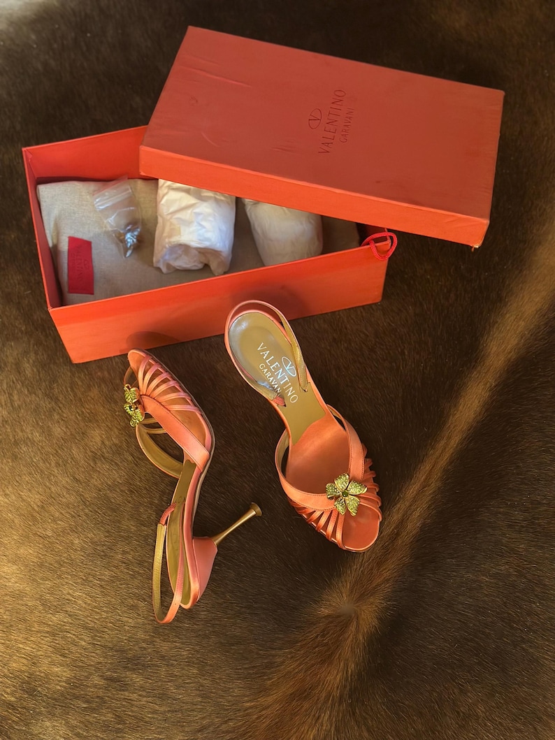 Puede incluir: Un par de sandalias de tac&oacute;n alto Valentino Garavani de color coral con un adorno floral. Se muestran junto a una caja coral abierta con el logotipo de Valentino Garavani. La caja contiene papel de seda y accesorios.