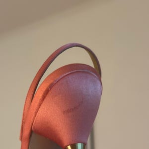 Puede incluir: Primer plano de un tac&oacute;n de sat&eacute;n rosa con un tac&oacute;n dorado. El zapato tiene una correa fina y un tac&oacute;n ligeramente curvado. El material de sat&eacute;n tiene un brillo sutil.