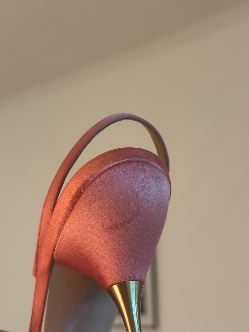 Puede incluir: Primer plano de un tac&oacute;n de sat&eacute;n rosa con un tac&oacute;n dorado. El zapato tiene una correa fina y un tac&oacute;n ligeramente curvado. El material de sat&eacute;n tiene un brillo sutil.