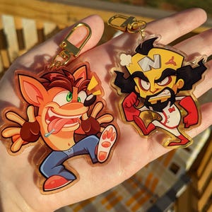 Puede incluir: Dos llaveros de personajes de dibujos animados. Uno presenta una criatura naranja con forma de zorro con un ojo verde, vaqueros azules y una lengua larga. El otro es un personaje con cabeza amarilla, pelo negro y un atuendo rojo y blanco. Ambos tienen cierres dorados.