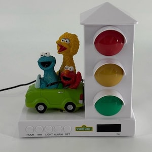 May include: A white Sesame Street alarm clock featuring Cookie Monster, Big Bird, and Elmo in a green car. The clock has a traffic light design with red, yellow, and green lights. The clock face has buttons and a digital display.