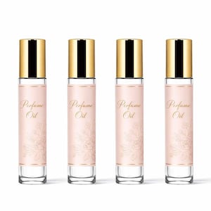 Op de afbeelding: Vier parfumolieflessen met gouden doppen en heldere glazen lichamen. Elke fles heeft een roze etiket met gouden tekst "Perfume Oil" en een bloemenontwerp. De flessen zijn in een rij gerangschikt.