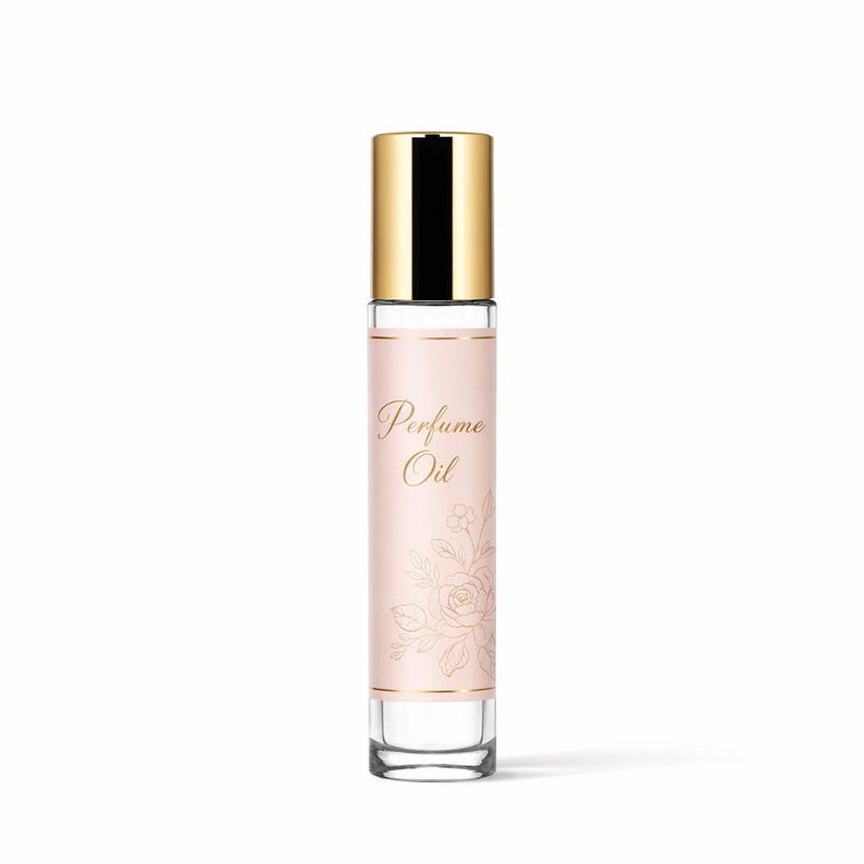 Op de afbeelding: Een parfumolie flesje met een gouden dop en een lichtroze etiket. Het etiket bevat de woorden "Perfume Oil" in gouden letters en een bloemenontwerp. De heldere glazen fles is cilindrisch.