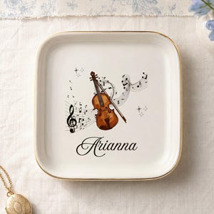 May include: A square white ceramic trinket dish with a gold-toned rim. The center features a brown violin illustration, black musical notes, a treble clef, and the name Arianna written in a black cursive script font on the surface.