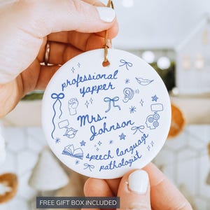 May include: A round white ceramic ornament featuring blue ink illustrations and text that reads professional yapper, Mrs. Johnson, and speech language pathologist, accented with small drawings of bows, stars, lips, an ear, and a brain.