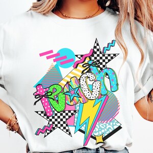 May include: White t-shirt with a colorful graphic design. The design features the word "Beach" in a playful font, surrounded by stars, lightning bolts, and geometric shapes in vibrant colors like pink, blue, and yellow.