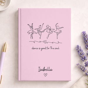 May include: A light pink hardcover journal featuring a black line art illustration of four ballet dancers, a small heart, and the text "dance is good for the soul". The name "Isabella" is printed below, followed by a small heart icon.