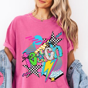 Puede incluir: Una camiseta rosa brillante con un dise&ntilde;o gr&aacute;fico colorido. El dise&ntilde;o incluye la palabra "Teach" en varias fuentes y patrones, junto con estrellas, rayos y formas geom&eacute;tricas. Las mangas est&aacute;n dobladas.