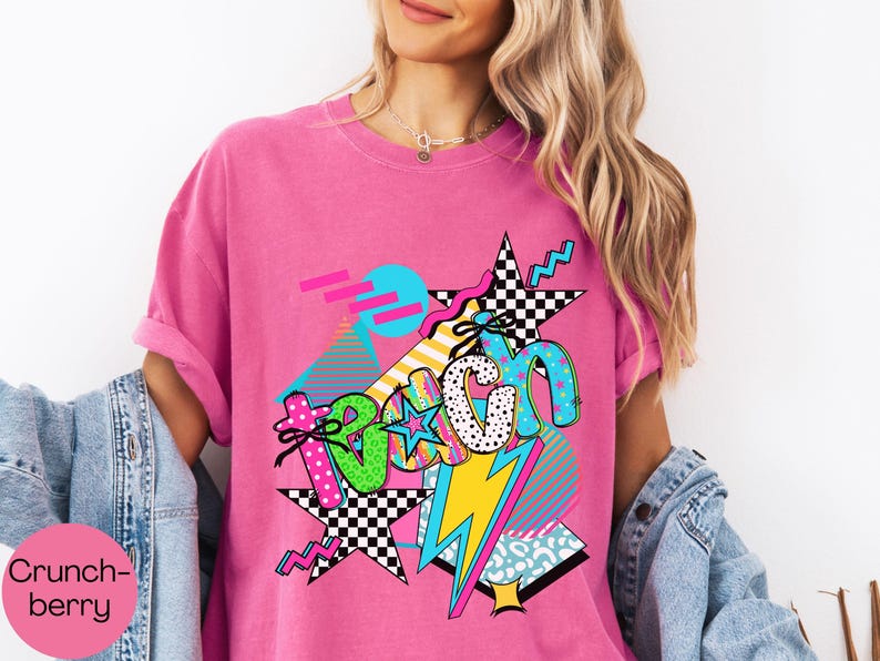 Puede incluir: Una camiseta rosa brillante con un dise&ntilde;o gr&aacute;fico colorido. El dise&ntilde;o incluye la palabra "Teach" en varias fuentes y patrones, junto con estrellas, rayos y formas geom&eacute;tricas. Las mangas est&aacute;n dobladas.
