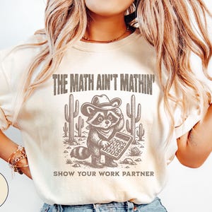 May include: Ivory-colored t-shirt with a graphic of a raccoon wearing a cowboy hat and holding a calculator. The text reads "THE MATH AIN'T MATHIN'" and "SHOW YOUR WORK PARTNER". The design includes cacti.