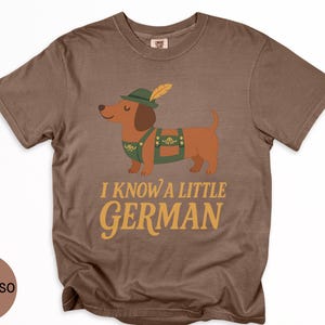 May include: Espresso-colored t-shirt featuring a cartoon dachshund wearing a green hat with a feather and green overalls. The text "I KNOW A LITTLE GERMAN" is printed in gold lettering below the dog.