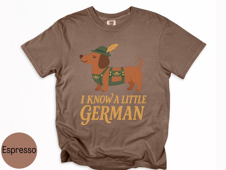 May include: Espresso-colored t-shirt featuring a cartoon dachshund wearing a green hat with a feather and green overalls. The text "I KNOW A LITTLE GERMAN" is printed in gold lettering below the dog.